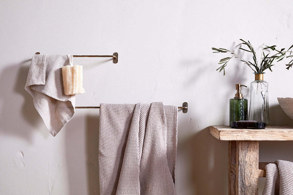 A minimal bathroom scene featuring beige towels on the nkuku Olani Bathroom Hanging Rail in Antique Brass, a loofah, and a rustic wooden shelf with a green glass soap dispenser, olive branches in a vase, and a bowl against a light wall.