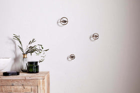 A minimalist scene features a light wooden table with a green glass jar, white bowl, black object, and clear vase with olive branches. On the plain white wall above are three Olani Bathroom Hooks in antique brass by nkuku.