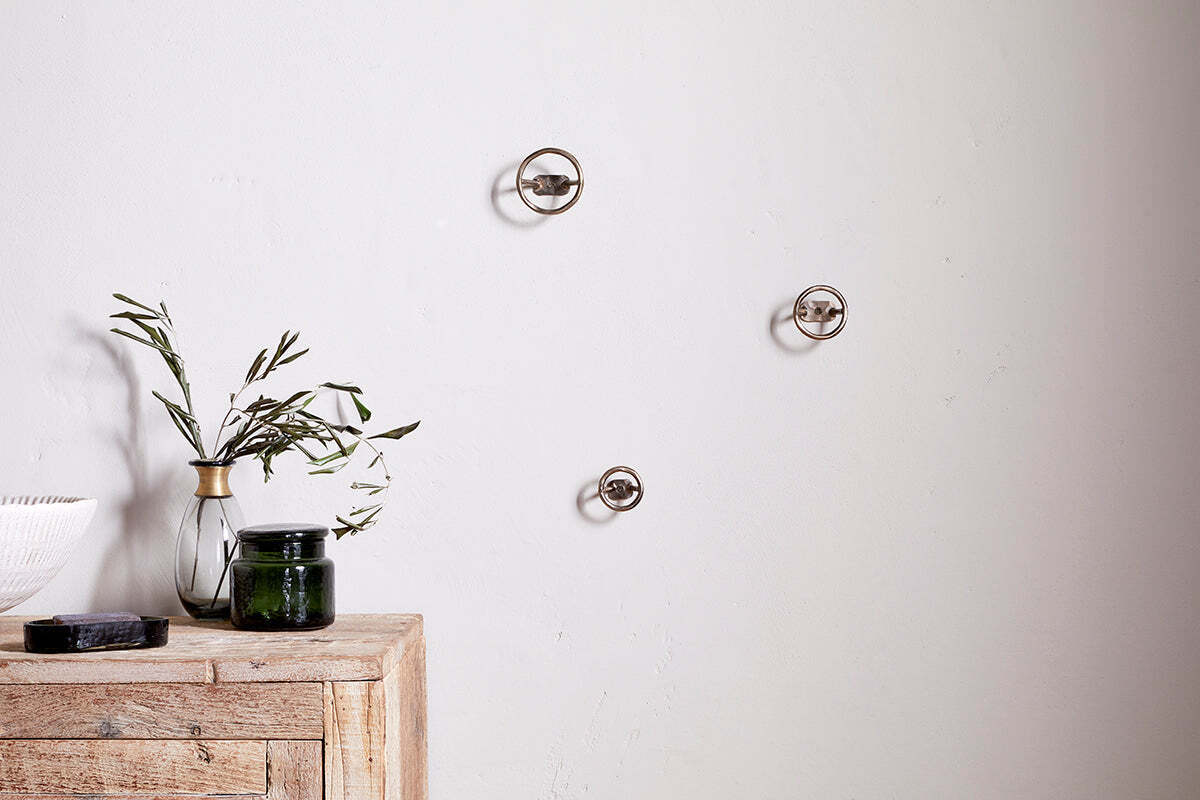 A minimalist scene features a light wooden table with a green glass jar, white bowl, black object, and clear vase with olive branches. On the plain white wall above are three Olani Bathroom Hooks in antique brass by nkuku.