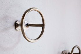 A close-up of an nkuku Olani Bathroom Hook in antique brass, featuring a round design with a horizontal bar, mounted on a white textured wall. A second hook from the set of 3 appears in the background for a spa-like feel.