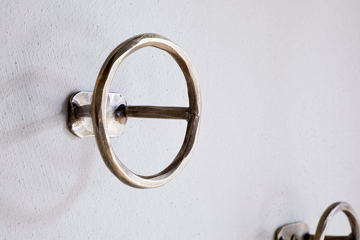 A close-up of an nkuku Olani Bathroom Hook in antique brass, featuring a round design with a horizontal bar, mounted on a white textured wall. A second hook from the set of 3 appears in the background for a spa-like feel.