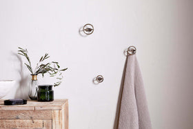 A minimalist spa bathroom features a light wooden cabinet, green branches in a vase, a dark candle, and a gray towel hanging on nkuku's Olani Bathroom Hooks in Antique Brass (Set of 3) against a plain white wall.