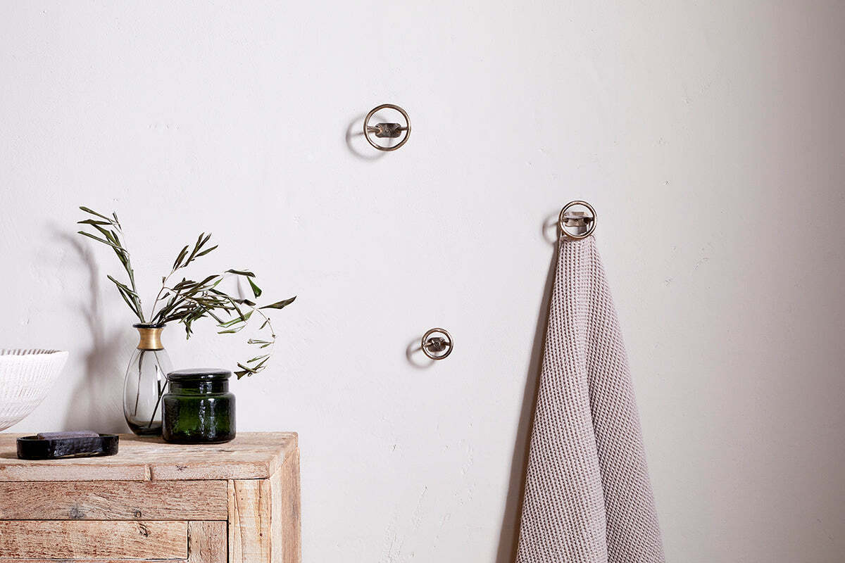 A minimalist spa bathroom features a light wooden cabinet, green branches in a vase, a dark candle, and a gray towel hanging on nkuku's Olani Bathroom Hooks in Antique Brass (Set of 3) against a plain white wall.