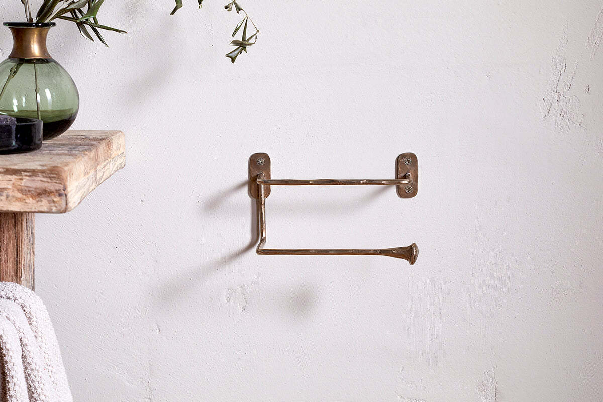 A brass double-arm towel bar is mounted on a white bathroom wall beside a wooden shelf with a green glass vase of leafy stems. Nearby, the nkuku Olani Toilet Roll Holder in Antique Brass complements the rustic, minimalist aesthetic.