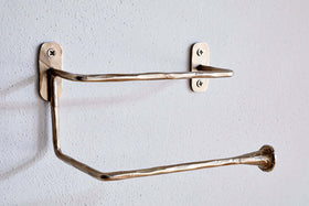 The Olani Toilet Roll Holder by nkuku features a handcrafted, hammered texture in antique brass. Its angular, stainless steel design mounts to a white bathroom wall with two visible screws per plate and stands empty, ready for a new roll.