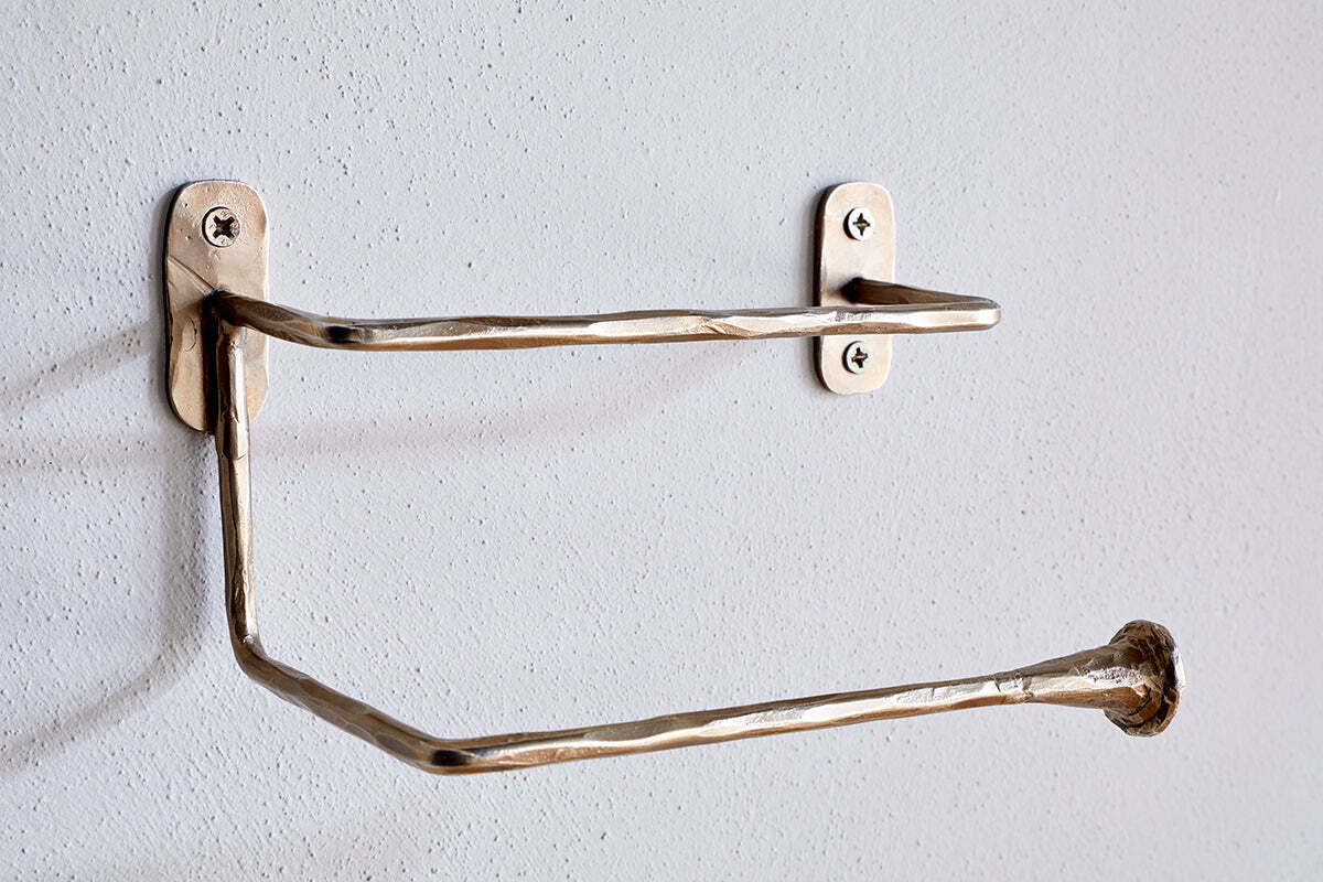The Olani Toilet Roll Holder by nkuku features a handcrafted, hammered texture in antique brass. Its angular, stainless steel design mounts to a white bathroom wall with two visible screws per plate and stands empty, ready for a new roll.
