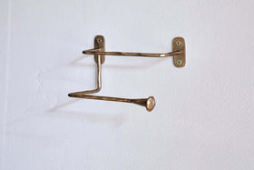 The nkuku Olani Toilet Roll Holder in antique brass features two bent, uneven bars with flared flat ends, hand-forged for a unique look. Wall-mounted with two screws, it casts a subtle shadow against the white bathroom wall.