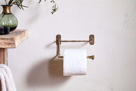 The Olani Toilet Roll Holder in antique brass by nkuku is mounted on a light bathroom wall with a toilet paper roll, beside a wooden shelf displaying a green vase and towel, enhancing the rustic, minimalistic decor.