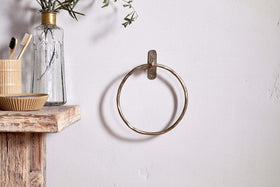 The Olani Towel Ring by nkuku in antique brass is mounted on a white wall. A rustic wooden shelf nearby holds a striped cup with toothbrushes, a small bowl, and a glass bottle with leafy stems for a minimal, natural bathroom look.