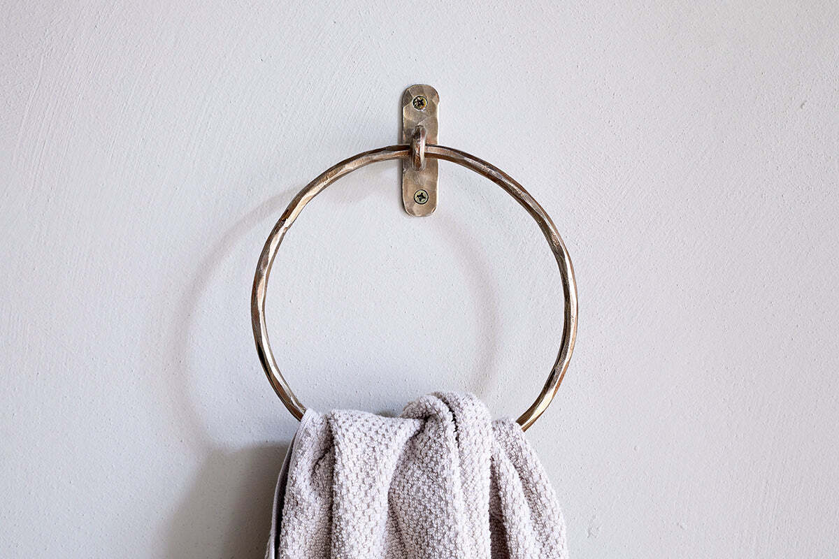 An Olani Towel Ring - Antique Brass by nkuku is mounted on a plain white wall, with a textured light gray hand towel draped over it, adding an elegant and minimal touch to the bathroom decor.