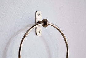 A close-up of the Olani Towel Ring - Antique Brass by nkuku, showing its textured, hammered surface and oval backplate with two visible screws, mounted on a pale grey wall as an elegant bathroom accessory.