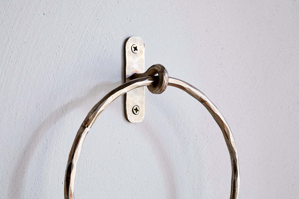 A close-up of the Olani Towel Ring - Antique Brass by nkuku, showing its textured, hammered surface and oval backplate with two visible screws, mounted on a pale grey wall as an elegant bathroom accessory.