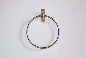 The Olani Towel Ring - Antique Brass by nkuku, a round bathroom accessory with a slightly twisted texture, is mounted on a plain white wall using a small metal bracket and two visible screws, with no towels hanging from it.