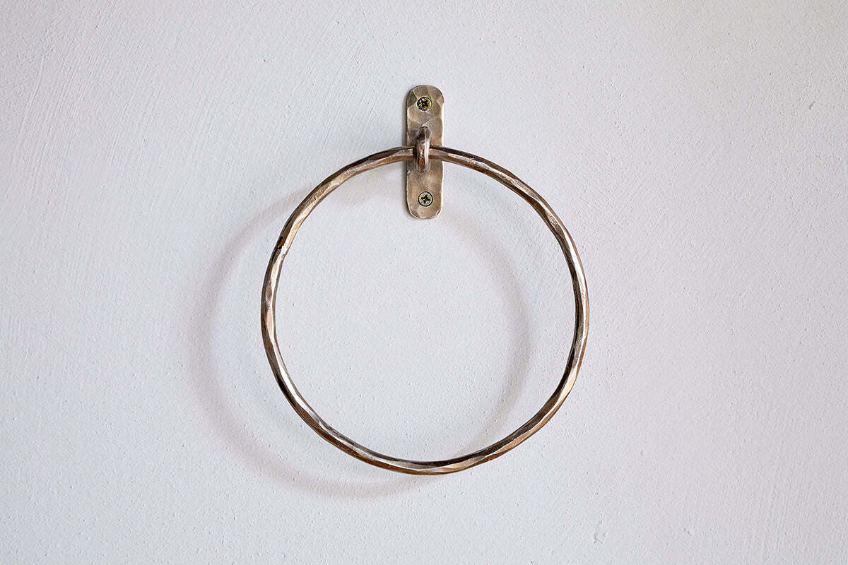 The Olani Towel Ring - Antique Brass by nkuku, a round bathroom accessory with a slightly twisted texture, is mounted on a plain white wall using a small metal bracket and two visible screws, with no towels hanging from it.