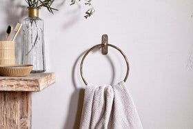An Olani Towel Ring - Antique Brass by nkuku, holding a textured white towel, is mounted on a white wall. Nearby rustic shelves display olive branches, toothbrushes, and a dish for a minimalist bathroom look highlighted by this sleek accessory.