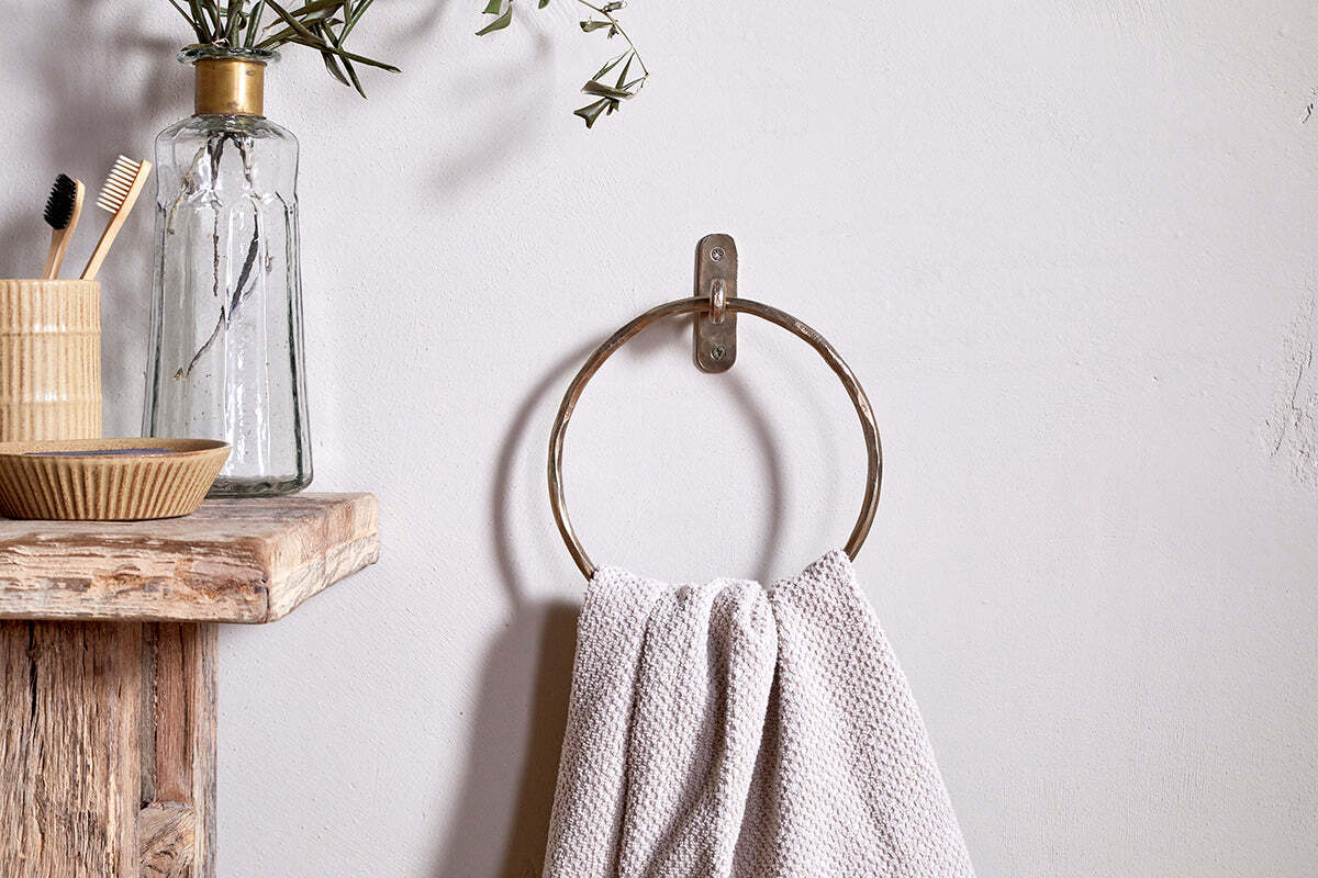 An Olani Towel Ring - Antique Brass by nkuku, holding a textured white towel, is mounted on a white wall. Nearby rustic shelves display olive branches, toothbrushes, and a dish for a minimalist bathroom look highlighted by this sleek accessory.