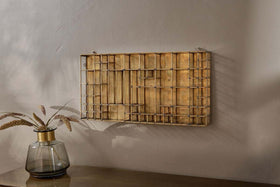 The Oni Printers Drawer - Brass by nkuku, a geometric gold wall sculpture inspired by traditional printing drawers, hangs on a light gray wall above wood. To the left, a gold-rimmed glass vase with wheat stems complements this unique wall art.