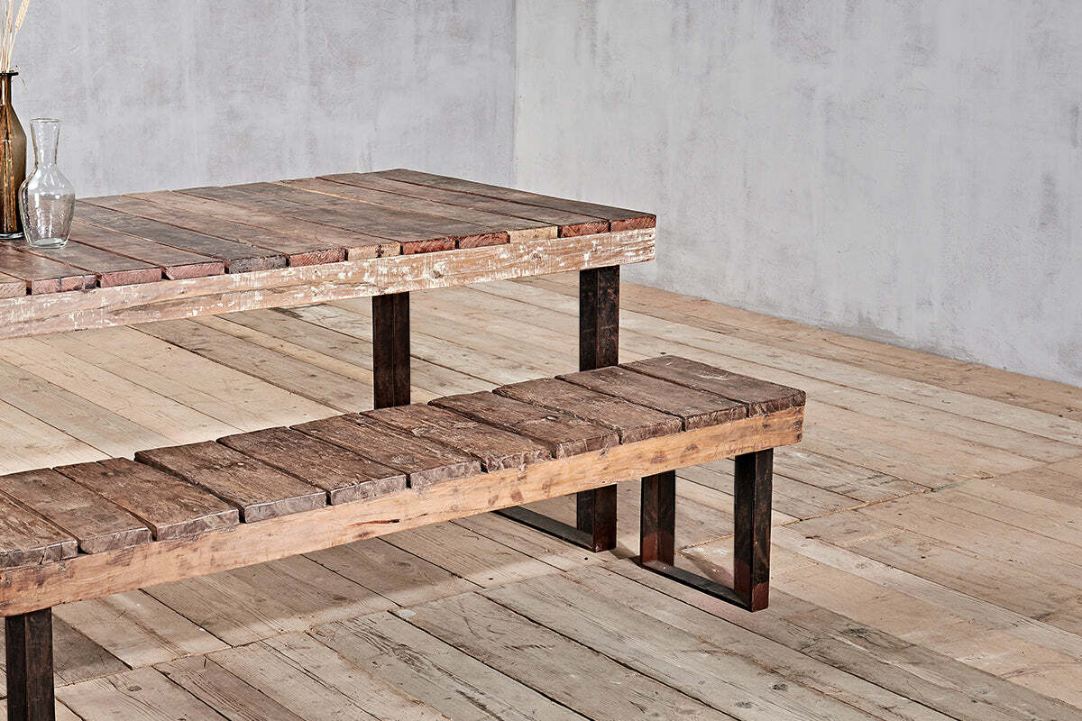 The nkuku Oso Wooden Bench, featuring rustic reclaimed wood and dark metal legs, sits next to a matching wooden table on a light floor against a plain gray wall. A glass bottle with dried plants decorates the table's corner.