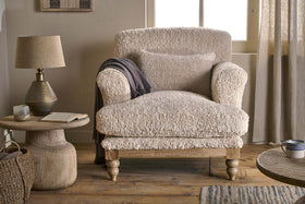 The nkuku Otas Teddy Cotton Armchair in Off White, paired with a matching pillow and draped with a dark gray throw, sits next to a round wooden side table holding a lamp, candle, and open book in a softly lit, neutral-toned room.