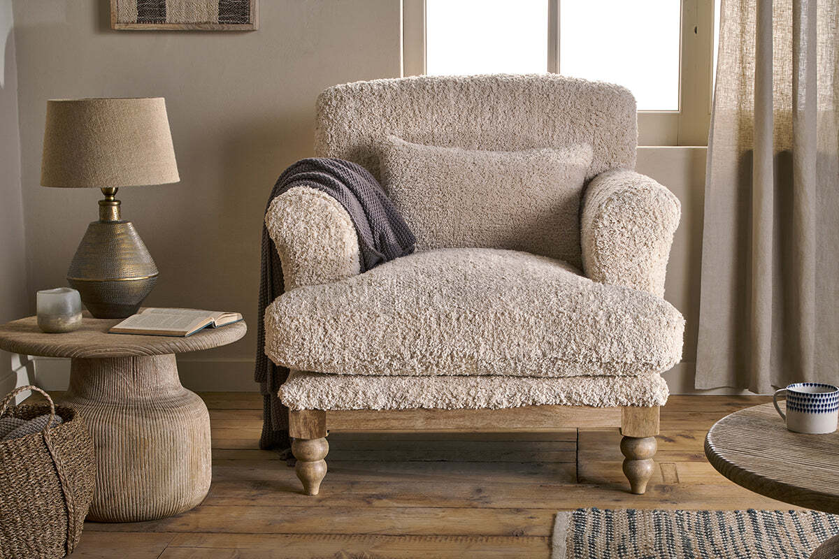 The nkuku Otas Teddy Cotton Armchair in Off White, paired with a matching pillow and draped with a dark gray throw, sits next to a round wooden side table holding a lamp, candle, and open book in a softly lit, neutral-toned room.