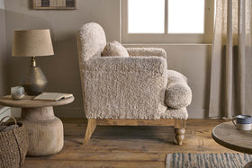 The Otas Teddy Cotton Armchair by nkuku in off white sits on a wooden floor beside a rustic side table featuring a lamp and an open book.