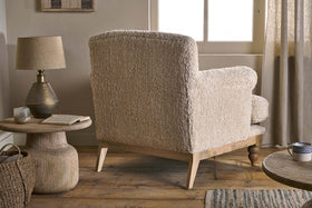 The Otas Teddy Cotton Armchair in off white by nkuku, with wooden legs, faces away from a sunlit window. To its left sits a round textured side table holding a lamp, book, glass, and woven basket on a wooden floor amid neutral decor.