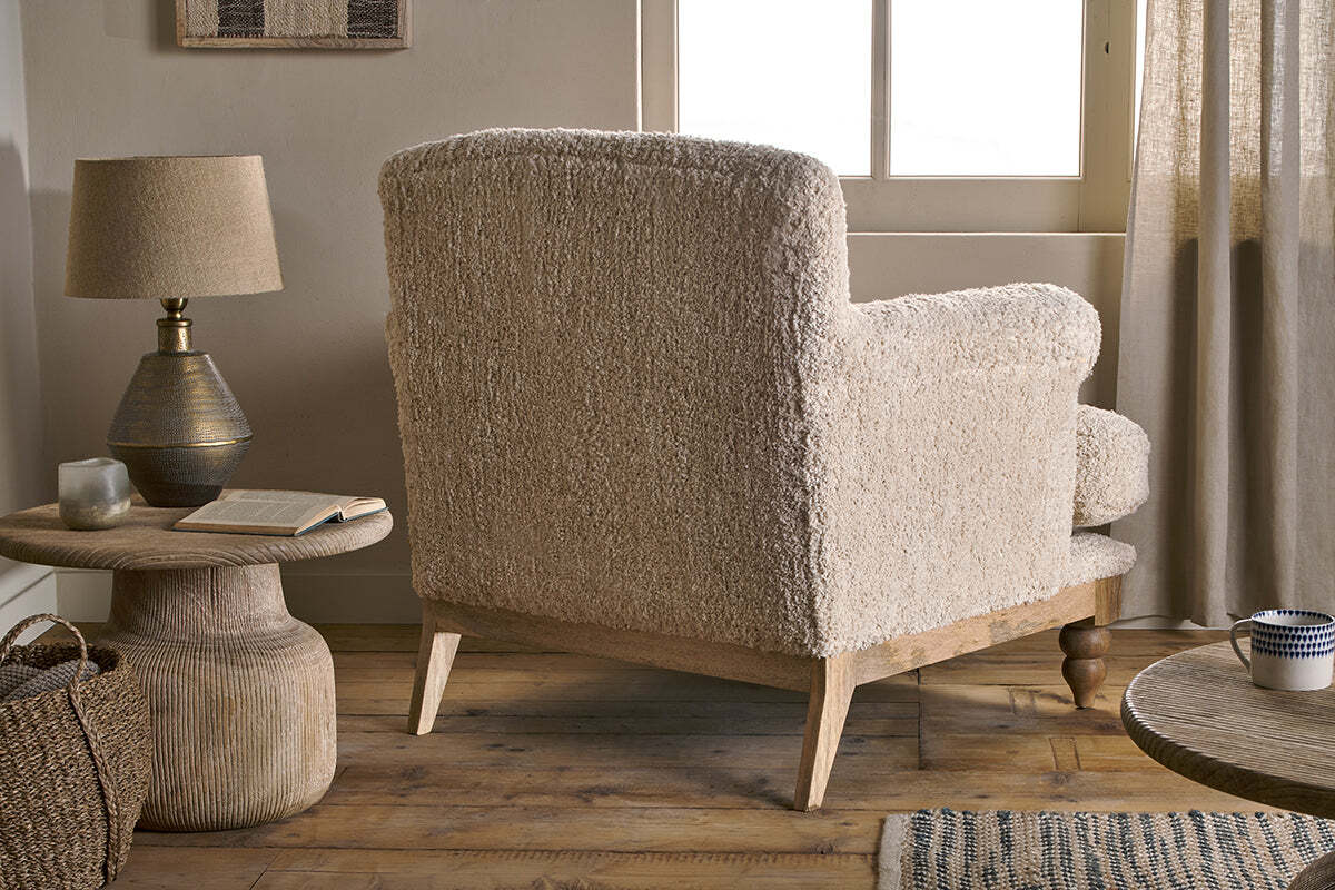 The Otas Teddy Cotton Armchair in off white by nkuku, with wooden legs, faces away from a sunlit window. To its left sits a round textured side table holding a lamp, book, glass, and woven basket on a wooden floor amid neutral decor.