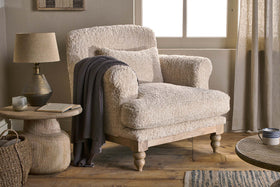 The Otas Teddy Cotton Armchair in Off White by nkuku, with wooden legs, is paired with a round mango wood side table holding a lamp, book, and candle. A dark knit blanket drapes over the chair as natural light warms the rustic room.