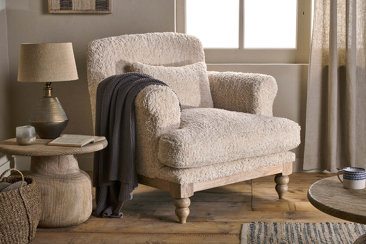 The Otas Teddy Cotton Armchair in Off White by nkuku, with wooden legs, is paired with a round mango wood side table holding a lamp, book, and candle. A dark knit blanket drapes over the chair as natural light warms the rustic room.