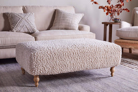 The Otas Teddy Ottoman in off white by nkuku, featuring wooden legs, stands on a woven rug before a cream sofa with textured pillows; a side table holding a mug and autumn leaves completes the cozy, neutral decor.