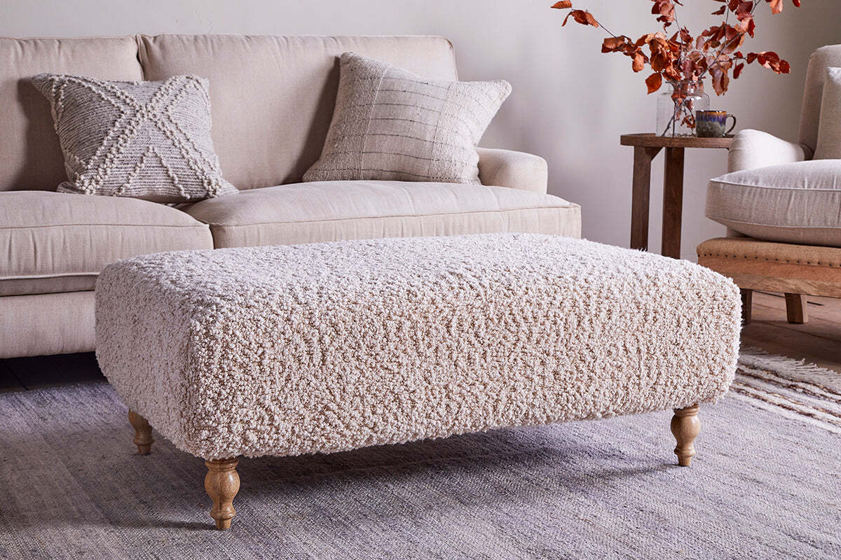 The Otas Teddy Ottoman in off white by nkuku, featuring wooden legs, stands on a woven rug before a cream sofa with textured pillows; a side table holding a mug and autumn leaves completes the cozy, neutral decor.