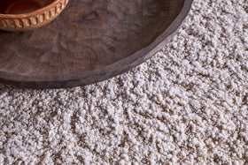 A close-up, angled shot shows a dark wooden tray and ceramic bowl resting on the nkuku Otas Teddy Ottoman - Off White, which features a mango wood base atop a plush, cream-colored shag rug.