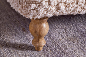 Close-up of a turned oak leg beneath nkuku’s Otas Teddy Ottoman - Off White. The cream boucle upholstery and textured grey rug accentuate the ottoman’s elegant, rounded form.