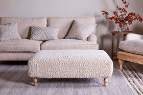A cozy living room with a beige sofa, three textured pillows, a large nkuku Otas Teddy Ottoman in off white with wooden legs, a neutral rug, and a side table holding a glass vase of autumnal branches and books.