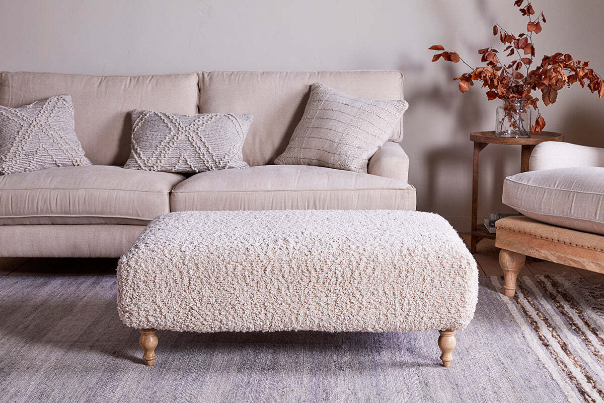 A cozy living room with a beige sofa, three textured pillows, a large nkuku Otas Teddy Ottoman in off white with wooden legs, a neutral rug, and a side table holding a glass vase of autumnal branches and books.