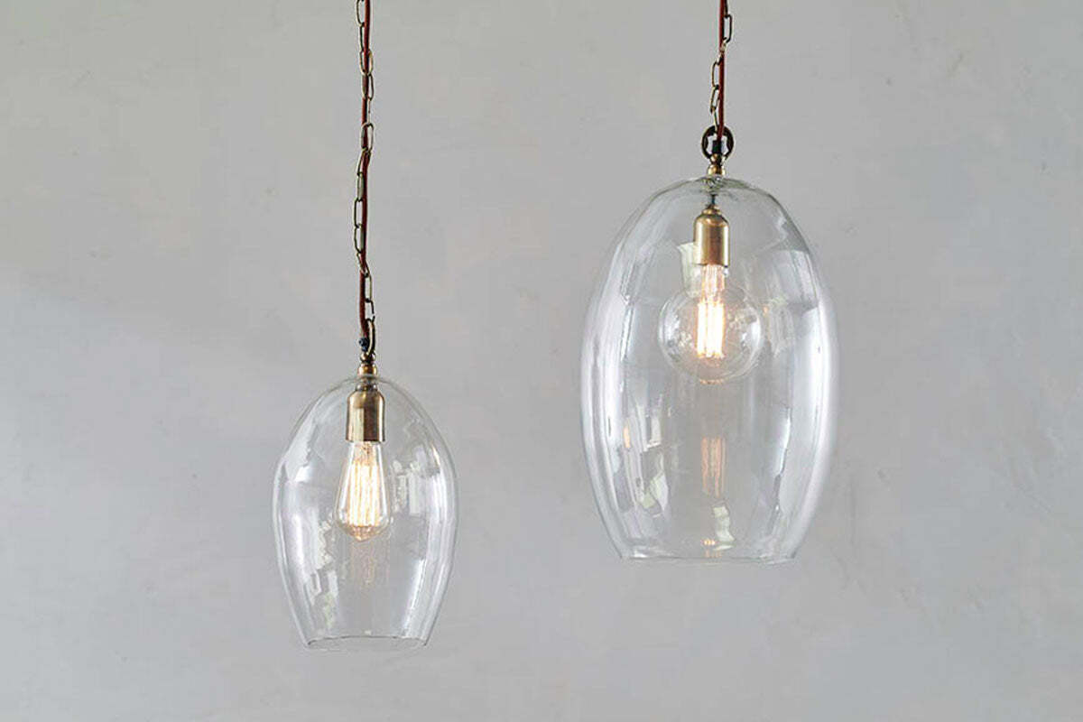 Two Otoro Recycled Glass Pendant - Clear - Small Oval lights by nkuku, with brass fittings and brown chains, hang against a light grey wall. The smaller pendant emits a warm glow, highlighting the bulbs and elegant recycled glass domes.