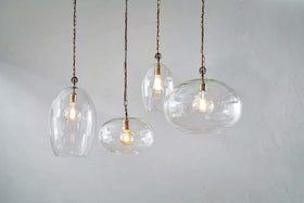 Four nkuku Otoro Recycled Glass Pendants in Clear Small Oval hang at varying heights with exposed bulbs and brass fittings against a light gray wall, each elegant glass shade showcasing a glowing bulb inside.