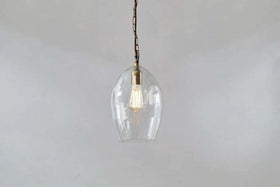 The Otoro Recycled Glass Pendant - Clear - Small Oval by nkuku features a dome-shaped glass shade and brass fittings with a vintage-style exposed bulb, making it a stylish, minimalist lighting choice.