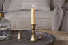 A lit cream-colored candle in the nkuku Prema Candle Holder - Antique Brass sits on a dark wooden tray beside a candle snuffer, with a glass vase and a beige sofa featuring textured pillows and a blanket in the background.