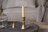 A cream candle in the nkuku Prema Candle Holder - Antique Brass sits on a dark round tray with a candle snuffer. In the background, a beige couch with textured pillows and a blanket is visible.