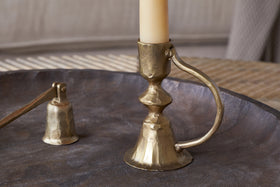 The nkuku Prema Candle Holder in antique brass, paired with a cream candle and matching snuffer, is displayed on a round metal tray against a softly blurred backdrop.