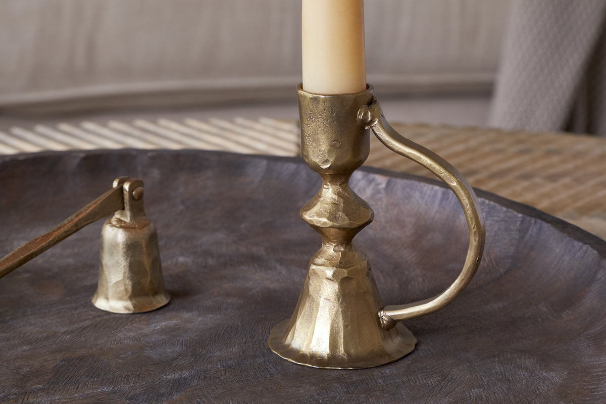 The nkuku Prema Candle Holder in antique brass, paired with a cream candle and matching snuffer, is displayed on a round metal tray against a softly blurred backdrop.