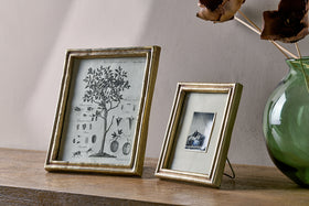Displayed on a wooden surface are two nkuku Pambani Photo Frames in antique brass: one with a vintage botanical print, the other showcasing a black and white mountain photo. A green vase with dried brown flowers sits to the right.