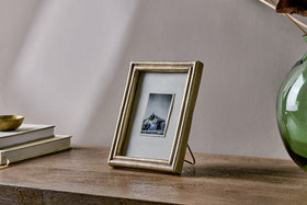 A black-and-white photo in the nkuku Pambani Photo Frame – Antique Brass sits on a wooden table with stacked books and a green glass vase beside it, set against a light-colored plain wall.
