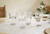 The nkuku Pani Highball - Clear (Set of 4) recycled glass tumblers are filled with water on a light tablecloth, accompanied by woven placemats, two wooden chairs, and a glass vase with eucalyptus—ideal for everyday dining.