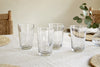 The nkuku Pani Highball - Clear (Set of 4) glassware is displayed on a light tablecloth, paired with woven placemats, wooden chairs with woven seats, and a vase of eucalyptus for a relaxed dining vibe.