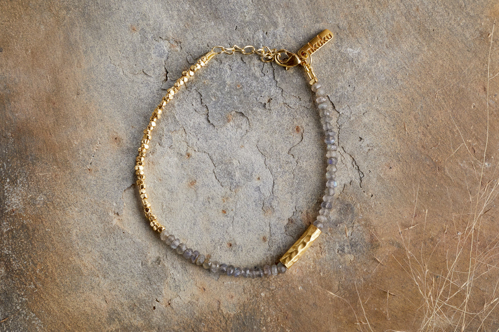The Perdu Labradorite Bracelet - Gold by nkuku features small gray labradorite beads, gold brass accents, a rectangular gold charm, and a gold chain clasp—a stunning natural stone accessory displayed on textured brown.