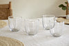 The nkuku Pani Tumbler - Clear (Set of 4), including recycled glass, are displayed on a light linen tablecloth with two woven placemats and a wooden chair with woven backrest. Green leaves decorate the upper right corner.