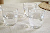The nkuku Pani Tumbler - Clear (Set of 4) is shown filled with water on a light fabric tablecloth, with a woven placemat in the background, highlighting their lightweight and durable recycled glass design.