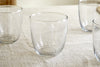 The nkuku Pani Tumbler - Clear (Set of 4) features four lightweight, durable recycled glass tumblers arranged on a light-textured fabric, with three near the edges and one centered in the image.